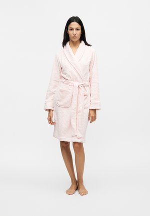 Femme debout pieds nus portant un peignoir matelassé rose clair noué à la taille avec une poche avant sur un fond blanc uni.