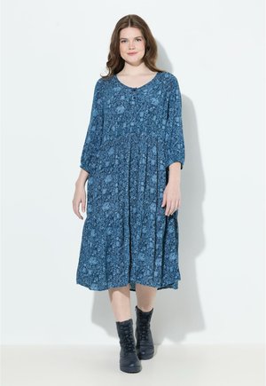 Blaues Blumendruckkleid mit rundem Halsausschnitt, dreiviertellangen Ärmeln und einem gerafften Rock. Getragen mit schwarzen Schnürstiefeln.