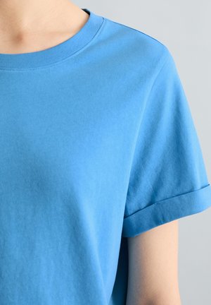 Persoon die een effen blauw T-shirt met korte mouwen en een ronde hals draagt tegen een neutrale grijze achtergrond.