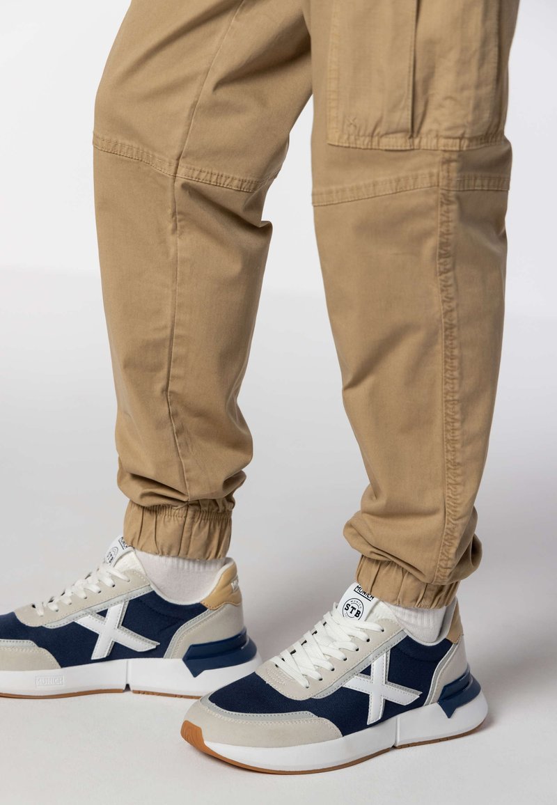 Beige hlače s elastičnim manžetama uparene s mornarsko plavim i bijelim tenisicama s teksturiranim gornjim dijelom, bijelim detaljima i potplatom od gume boje gume.