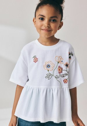 Junges Mädchen mit lockigem Haar, das ein weißes kurzärmeliges Oberteil mit bestickten bunten Blumen und blaue Jeans trägt, lächelt sanft.