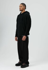 Schwarzer, strukturierter Fleece-Hoodie mit Kängurutasche und Kordelzugkapuze, kombiniert mit lockeren schwarzen Hosen und klobigen schwarzen Schuhen.