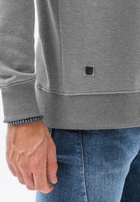 Grijze sweatshirt met ribgebreide manchetten en zoom, met een klein logo-label. Hand zichtbaar, met een blauwe kralenarmband. Jeans zijn donkerblauw.