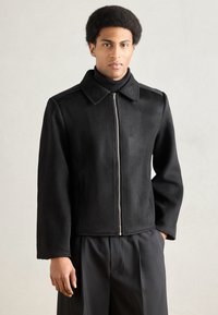 Crna jakna na patentni zatvara�č s oštrim ovratnikom, izrađena od meke tkanine. Ima bočne džepove i toniranu šavove. Nosi se preko crne rolke.