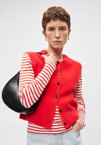 Κόκκινο ριμπ πλεκτό γιλέκο με μαύρα κουμπιά, φορεμένο πάνω από λευκή και κόκκινη ριγέ μακρυμάνικη μπλούζα. Μαύρη τσάντα κρεμασμένη στον ώμο.
