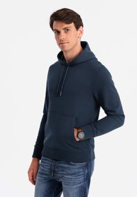Felpa blu navy in cotone, con tasca frontale, cappuccio regolabile e bordo a costine. Abbinata a jeans blu e a un orologio.