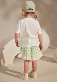 White short-sleeve T-shirt with a logo tag, paired with green checkered shorts featuring a lace trim. Worn with beige high-top shoes.