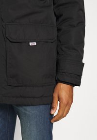 Veste matelassée noire avec une poche avant et un écusson logo. Dispose d'une fermeture éclair et de poignets cousus. Portée avec un jean bleu.