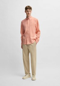 Camicia button-up a maniche lunghe di un rosa chiaro, in misto lino, abbinata a pantaloni beige e sneakers crema, in piedi davanti a uno sfondo neutro.