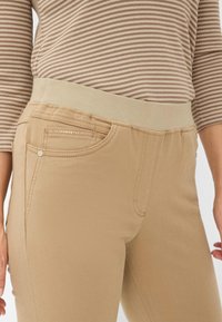 Beige broek met elastische tailleband en zijzakken, voorzien van een gladde stofstructuur en subtiele decoratieve stiksels op de voorzak.