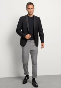 Veste de smoking noire avec revers en satin, portée sur une chemise bleu marine. Pantalon gris ajusté et chaussures en cuir noir complètent la tenue.