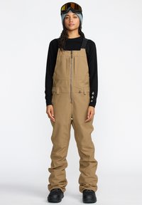 Salopette marron avec bretelles réglables, fermeture �éclair à l'avant et poches ; portée par-dessus un t-shirt à manches longues noir et associée à des lunettes de ski et des bottes.