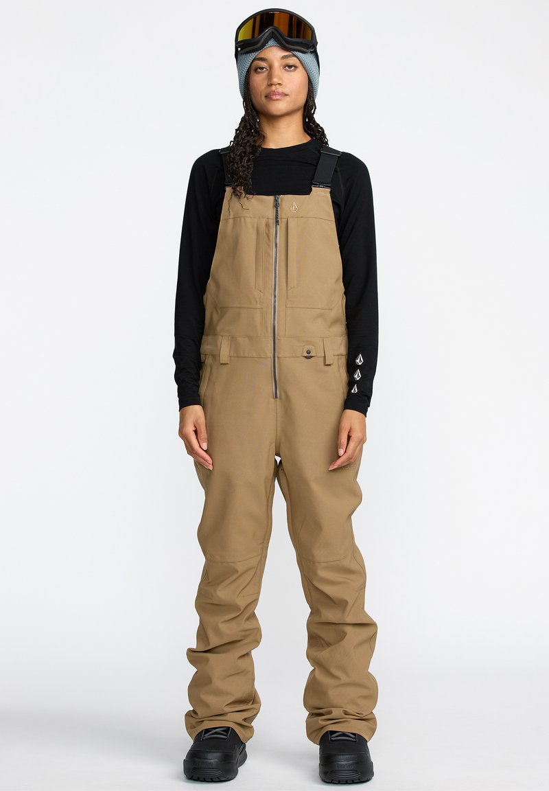 Salopette marron avec bretelles réglables, fermeture éclair à l'avant et poches ; portée par-dessus un t-shirt à manches longues noir et associée à des lunettes de ski et des bottes.