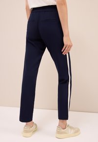 Persona che indossa pantaloni blu scuro con strisce laterali bianche e sneakers beige Veja, in piedi contro uno sfondo beige uniforme.