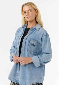 Jasnoniebieska koszula z denimu o lekko oversize'owym kroju, z zapięciem na guziki z przodu, dwiema kieszeniami na piersiach oraz podwiniętymi rękawami. Posiada wyblakłą teksturę.