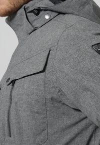 Graue Stoffjacke mit hohem Kragen, Reißverschluss und Druckknopfverschluss sowie seitlicher Tasche mit Klappe. Strukturierter Stoff mit einem dezenten Muster.