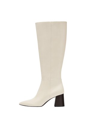 Botte en cuir couleur crème montant jusqu’au genou avec bout pointu et talon bloc foncé sur fond blanc.