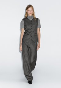 Gilet grigio su misura con chiusura a bottoni, abbinato a un dolcevita grigio a maniche corte e a pantaloni ampi grigi. Tessuto morbido, design minimalista.