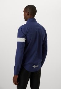 Marinefarbene Radjacke mit einem weißen Streifen am linken Ärmel, ausgestattet mit einem hohen Kragen und einem gestickten "Rapha"-Logo auf der Rückseite. Glatte Oberflächenstruktur.