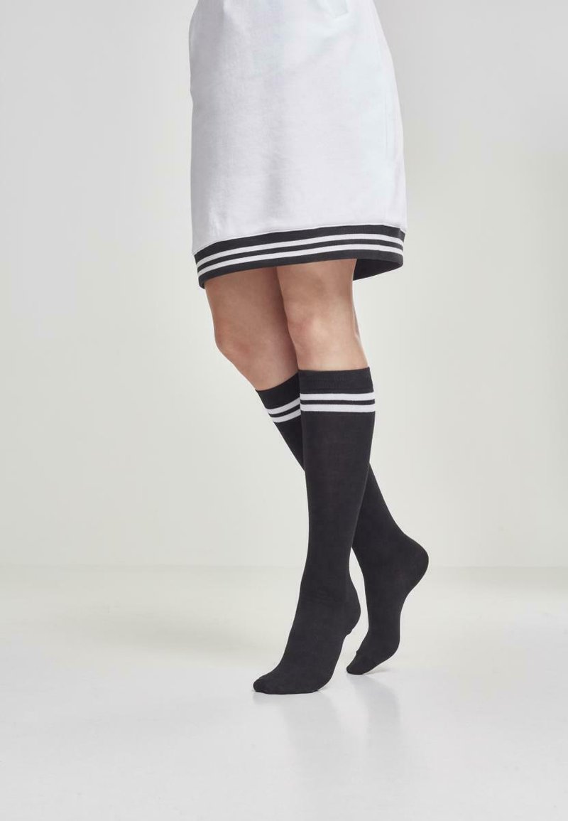 Calcetines negros hasta la rodilla fabricados con una tela suave, con rayas horizontales blancas en la parte superior, combinados con una falda blanca que presenta un dobladillo con rayas negras.