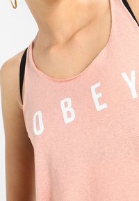 Ljusrosa linne i mjukt material, med vit text "OBEY." Bred halsringning och ärmhål, lager på lager över en svart topp under.