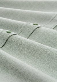 Světle zelená tkanina s vzorem rybí kost, obsahující dva knoflíky a jemnou strukturu, ukazující složené části a jemné detaily šití.