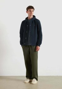 Giovane uomo che indossa una giacca con cappuccio blu navy, pantaloni larghi verde scuro, una camicia grigia e sneakers bianche, in piedi davanti a uno sfondo chiaro e neutro.