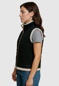 Zwart mouwloos gewatteerd vest met beige fleece trim, voorritssluiting, zijzakken en textielstof. Draagt over een grijs T-shirt met korte mouwen.