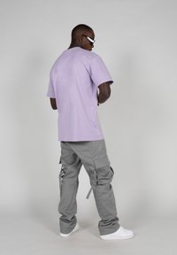 Tricou din bumbac violet și pantaloni cargo gri, cu multiple buzunare și bretele ajustabile, asortați cu adidași albi și ochelari de soare.