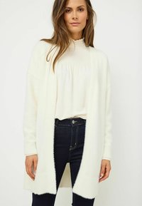 Biała, puszysta sweterka z dzianiny z odkrytym przodem, z prążkowanymi detalami przy mankietach i u dołu, w zestawieniu z kremową bluzką i ciemnymi dżinsami.