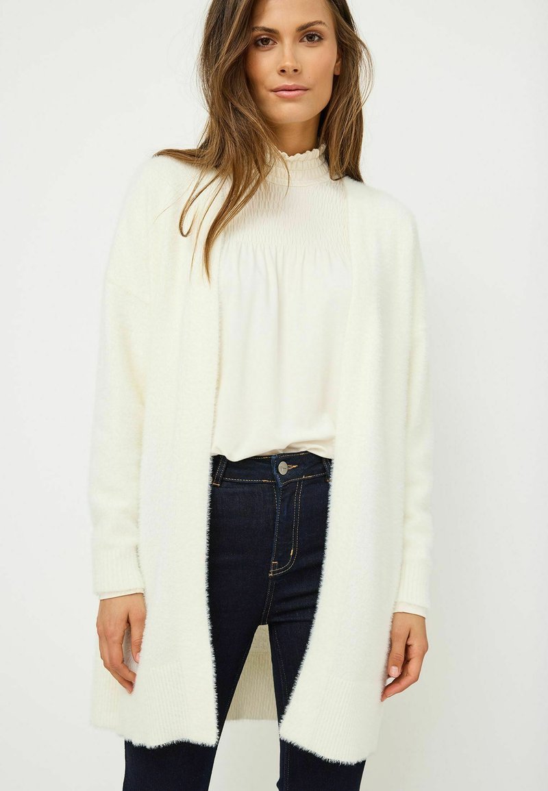 Biała, puszysta sweterka z dzianiny z odkrytym przodem, z prążkowanymi detalami przy mankietach i u dołu, w zestawieniu z kremową bluzką i ciemnymi dżinsami.