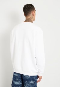 Valge dressipluus mugava lõikega, soonikkoes mansetid ja alläär. Sile puuvillane kangas, lihtne disain ilma logodeta, kantud siniste mustritega lühikeste pükstega.