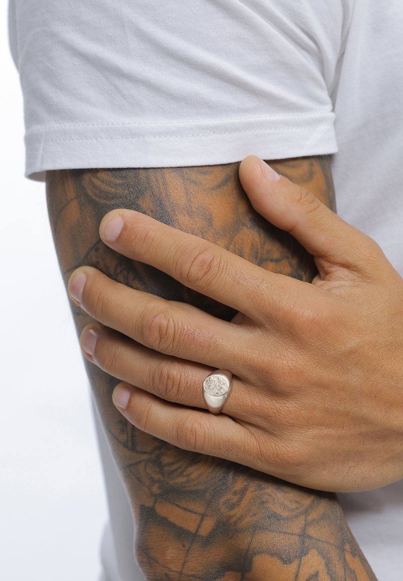 Hõbedane sõrmus ümarate õlgadega, millel on detailne graveering, kantakse käel, millel on nähtavad tätoveeringud, halli tausta ees.