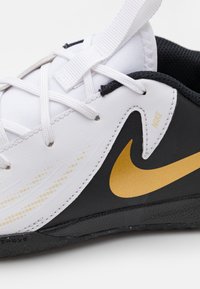 Witte en zwarte sneaker met een gouden Nike-swoosh, voorzien van een textuur, vetersluiting en zichtbare stiksels.