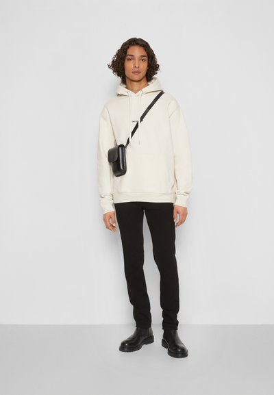 Hanorac cu glugă de culoare crem deschis, cu buzunar frontal, purtat cu pantaloni negri slim și botine negre până la gleznă. Include o geantă mică neagră tip crossbody.