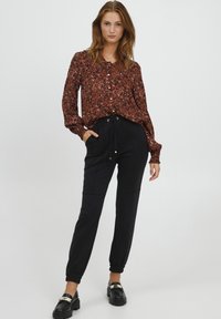 Geprinte blouse in aardetinten met een knoopsluiting aan de voorkant, gecombineerd met zwarte tapering broek met een elastieken tailleband en trekkoord. Zwarte instappers.