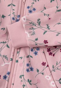 Vaaleanpunainen vetoketjullinen takki, jossa on kukkakuosi, jossa on sinisiä, purppuranvärisiä ja valkoisia kukkia; ribbit hihansuissa ovat yhteensopivaa vaaleanpunaista kangasta.
