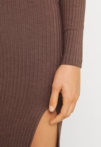Gros plan sur une main reposant près d'une fente haute jusqu'à la cuisse sur une robe marron à manches longues côtelée.