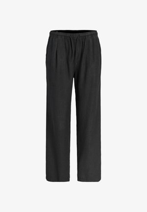 Tinejdžer s kovrčavom kosom stoji nasmijan, noseći bež košulju kratkih rukava s gumbima preko bijele majice, crne široke hlače i bijele tenisice.