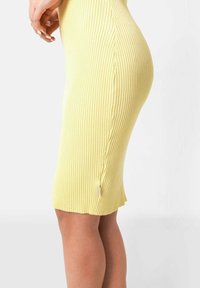Robe en tricot côtelé jaune avec une coupe ajustée, longueur genou et une fente sur le côté. Présente une texture douce et une étiquette de marque subtile près de l'ourlet.