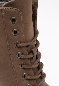 Brązowy skórzany but z metalowymi oczkami, starannie wykończonym przeszyciem i okrągłymi sznurówkami. Wnętrze zdobi fakturowana tkanina, co podnosi komfort użytkowania.