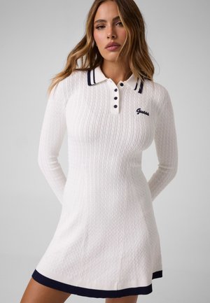Un mannequin porte une robe polo Guess blanche en maille torsadée, rehaussée de finitions bleu marine, et pose devant un fond de studio gris uni.