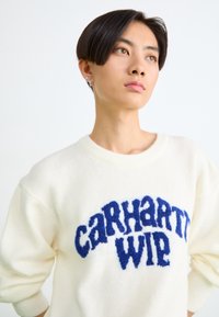 Kreemikas värvi kudum, millel on tumesinine "CARHARTT WIP" logo. Mugav lõige, soonikkrae ja pehme tekstuur.