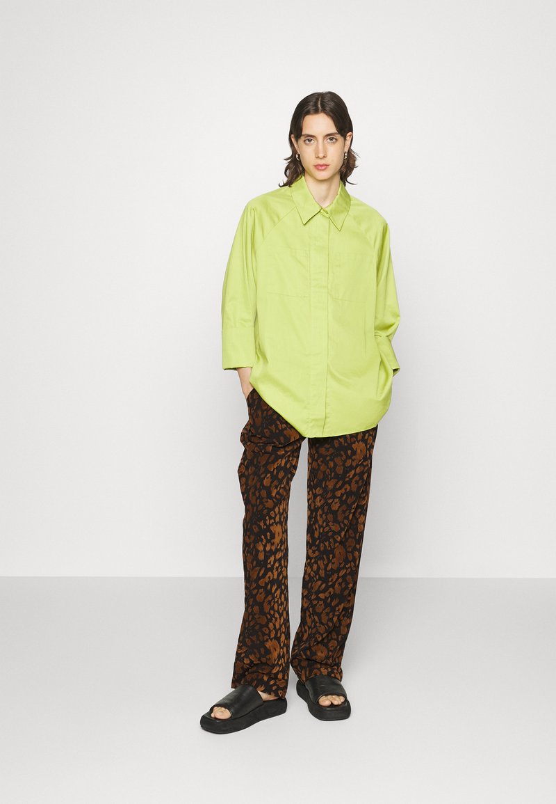 Hellgrünes, oversized Hemd mit spitzem Kragen, kombiniert mit dunkelbraunen Hosen im Tiermuster. Schwarze Rutschsandalen runden das Outfit ab.