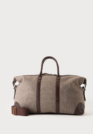 Grand sac fourre-tout en tissu beige avec poignées, sangles et base en cuir marron foncé, posé sur un fond clair uni.