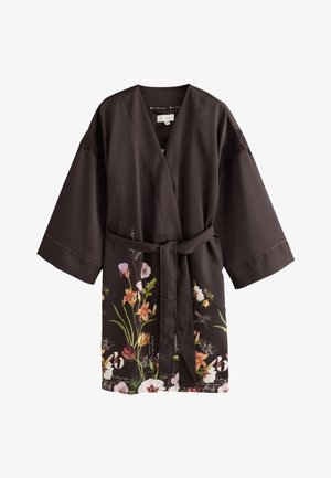 Kvinde står barfodet i en mørk blomsterkjole bundet ved taljen i et blødt oplyst soveværelse med neutral indretning og en blomstervase på et sidebord.