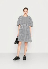 Vestido de padrão gingham preto e branco com mangas bufantes, cintura ajustada e saia evasê. Combinado com sapatos de plataforma pretos e uma pequena mala.