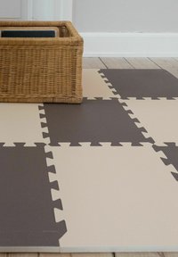 Πλεκτό καλάθι σε μπεζ και σκουρόχρωμα γκρι συνδεόμενα πλακάκια από αφρό σε δάπεδο, μπροστά σε λευκό τοίχο και ξύλινο δάπεδο.