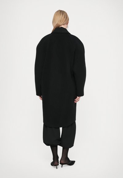 Fekete gyapjúkabát, amelynek oversized vállai, széles gallérja és hosszú ujjai vannak. Egyenes szabású, laza fekete nadrággal és magassarkúval párosítva.