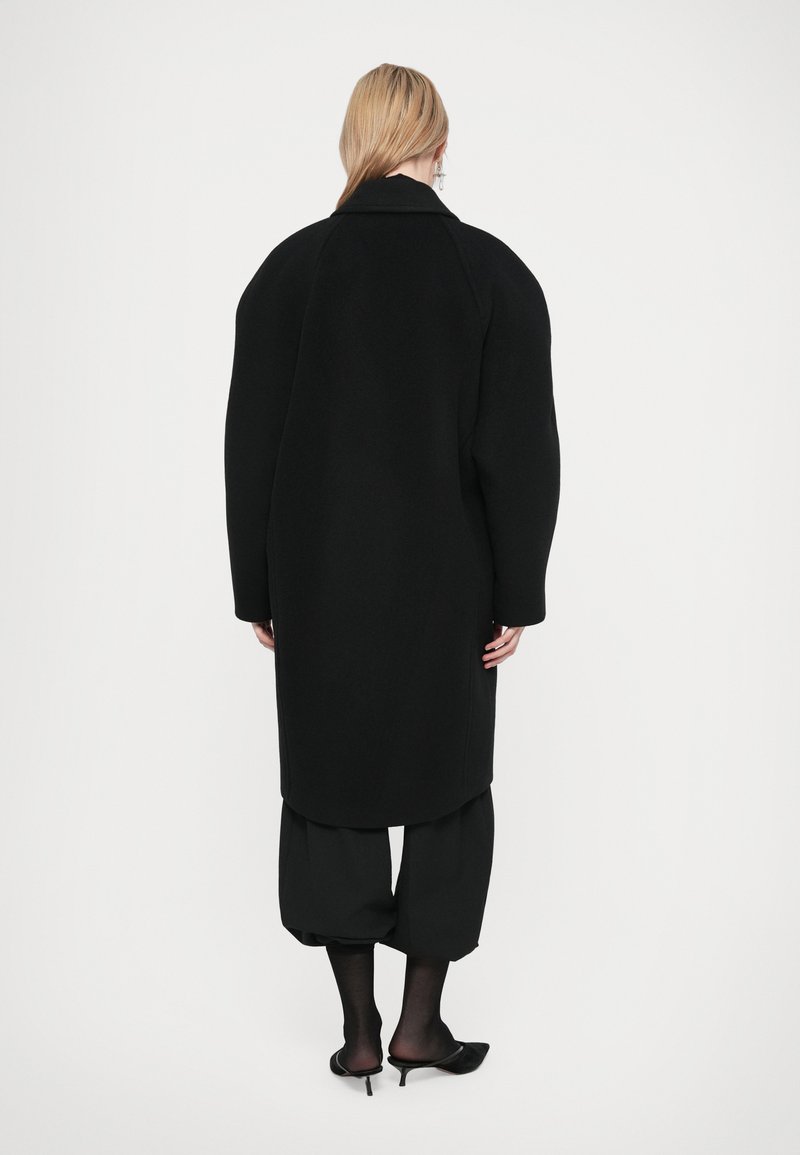 Svart ullrock med överdimensionerade axlar, en bred krage och långa ärmar. Har en rak skärning, kombinerad med lösa svarta byxor och klackar.